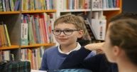 Chłopiec w jasnych włosach i okularach, ubrany w niebieski sweterek. Obok niego inne dzieci. Za nimi regał z książkami. 